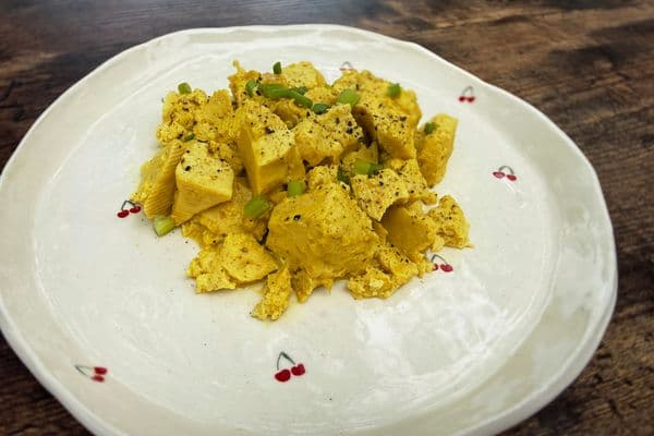 A golden plant-based tofu scramble on a white cherry patterned plate, seasoned with black pepper, plant-based egg sauce and topped with sliced green onions, served on a rustic wooden surface.