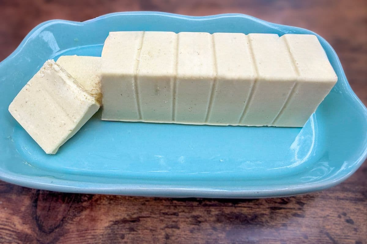 Side view of a homemade plant-based butter block with one slice removed on a light blue ceramic butter dish, showing the smooth dense texture of the vegan butter on a rustic wooden surface.