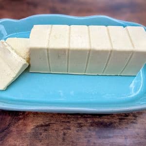 Side view of a homemade plant-based butter block with one slice removed on a light blue ceramic butter dish, showing the smooth dense texture of the vegan butter on a rustic wooden surface.