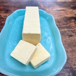 A block of homemade plant-based butter with two slices cut and laid flat on a light blue ceramic butter dish on a rustic wooden surface, showing the smooth creamy interior.