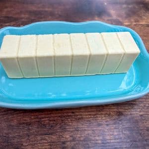 A full stick of homemade plant-based butter with pre-scored slices sitting on a light blue ceramic butter dish on a rustic wooden surface.