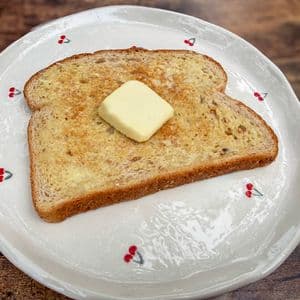 A slice of homemade plant-based butter melting on a slice of golden toasted multigrain bread on a white cherry patterned plate on a rustic wooden surface.