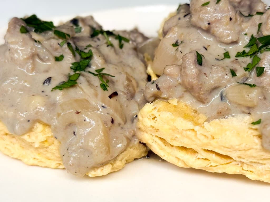 Close up of creamy plant-based beyond meat gravy smothering flaky vegan biscuits garnished with fresh chopped parsley showing the rich thick texture of the gravy.