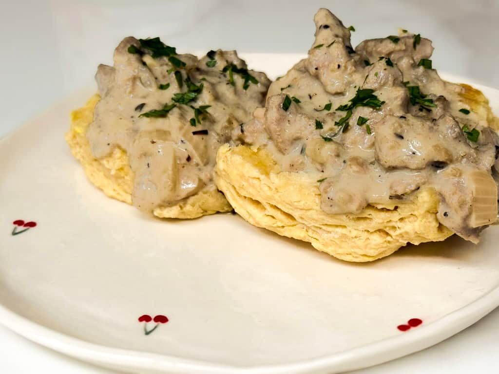 Two fluffy vegan biscuits topped with creamy plant-based beyond meat gravy garnished with fresh chopped parsley on a white cherry patterned plate.