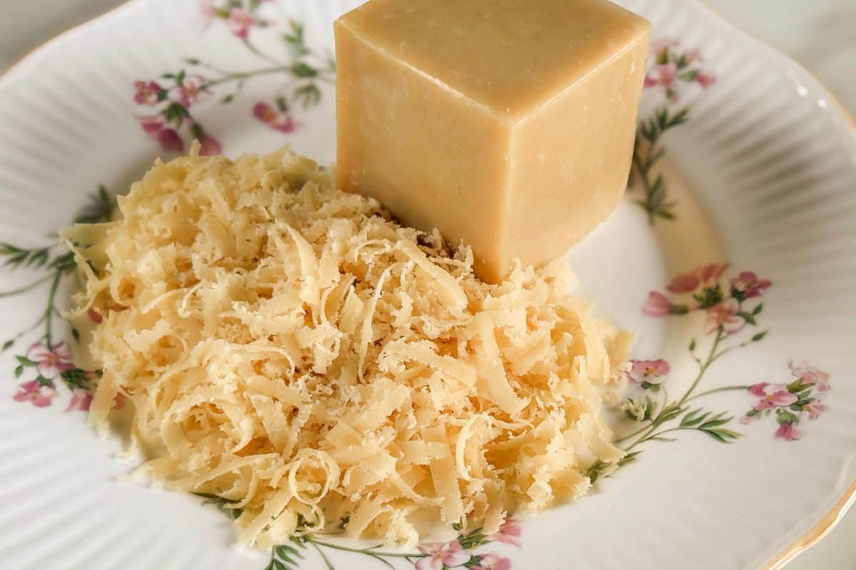 A block of homemade vegan parmesan cheese alongside a generous pile of freshly grated vegan parmesan, displayed on a vintage white ribbed ceramic plate with pink floral details and gold trim on a light background.