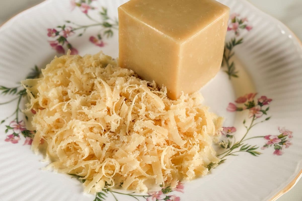 A block of homemade vegan parmesan cheese alongside a generous pile of freshly grated vegan parmesan, displayed on a vintage white ribbed ceramic plate with pink floral details and gold trim on a light background.