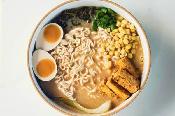 A bowl of vegan chicken paitan ramen with a rich creamy broth, curly ramen noodles, two halves of a plant-based soft-boiled egg with a golden yolk, crispy tofu, sweet corn, sliced green onions, broccolini, and bok choy, served in a white ceramic bowl with a tan rim on a marble surface.