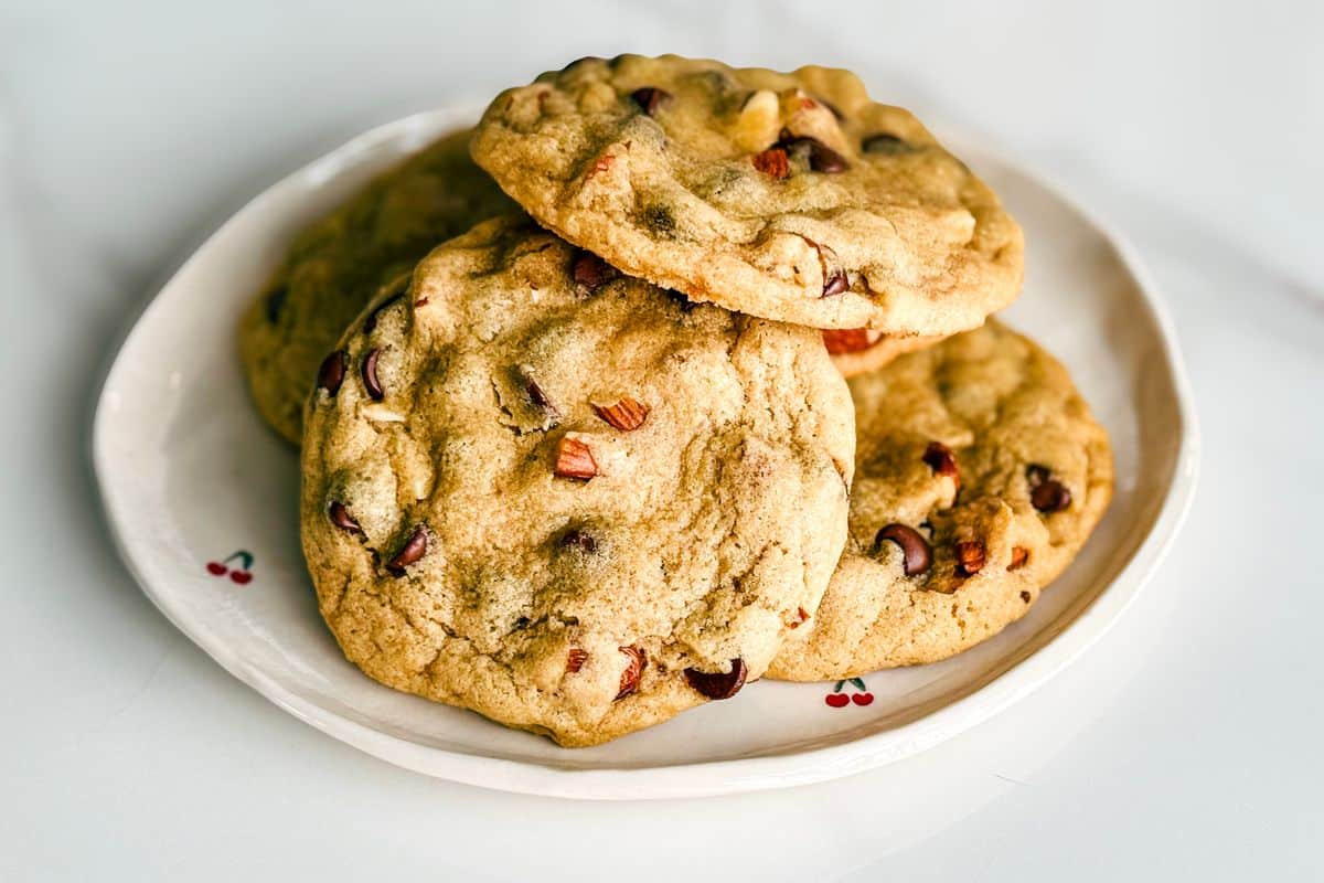 A plate of large, chewy chocolate chip cookies stacked on a white ceramic plate with cherry illustrations, photographed on a marble surface.
