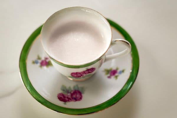 A smooth, pale vegan cream substitute served in a vintage white and green floral teacup with pink rose details and gold trim, resting on a matching saucer against a soft white background, photographed from above.