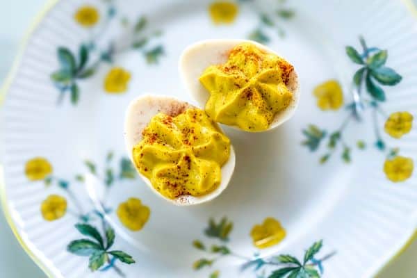 Two vegan deviled eggs made with tofu-based egg whites piped with a golden plant-based yolk filling and dusted with smoked paprika, displayed on a vintage white ceramic plate with a yellow rim and botanical floral details on a light background.