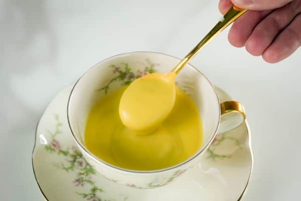 A hand holding a gold spoon lifting a scoop of rich, golden vegan hollandaise sauce from a vintage white floral teacup with gold trim on a matching saucer, photographed against a soft white background.