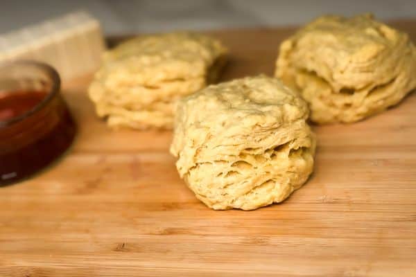 Three fluffy layered vegan biscuits on a bamboo cutting board with a small bowl of maple syrup and a block of plant-based butter blurred in the background.