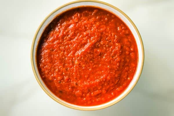 A bowl of rich, deeply red homemade vegan marinara sauce with a thick, chunky texture, served in a rustic white ceramic bowl with a tan rim against a soft light background.