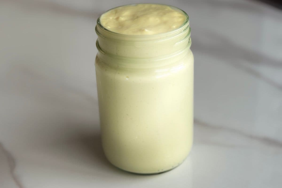 A glass jar filled with thick, pale yellow homemade mayonnaise, sitting on a white marble countertop.
