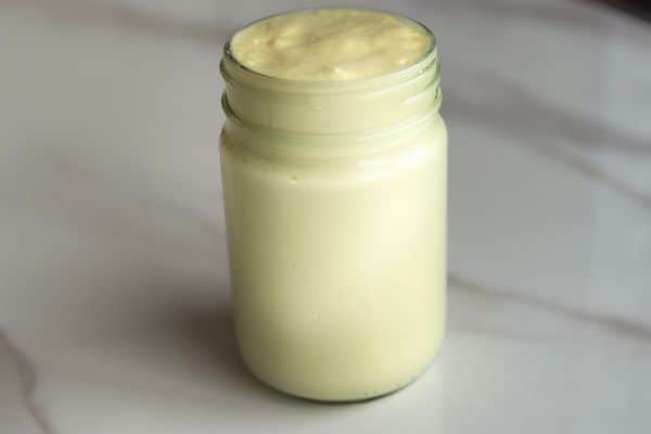 A glass jar filled with thick, pale yellow homemade mayonnaise, sitting on a white marble countertop.