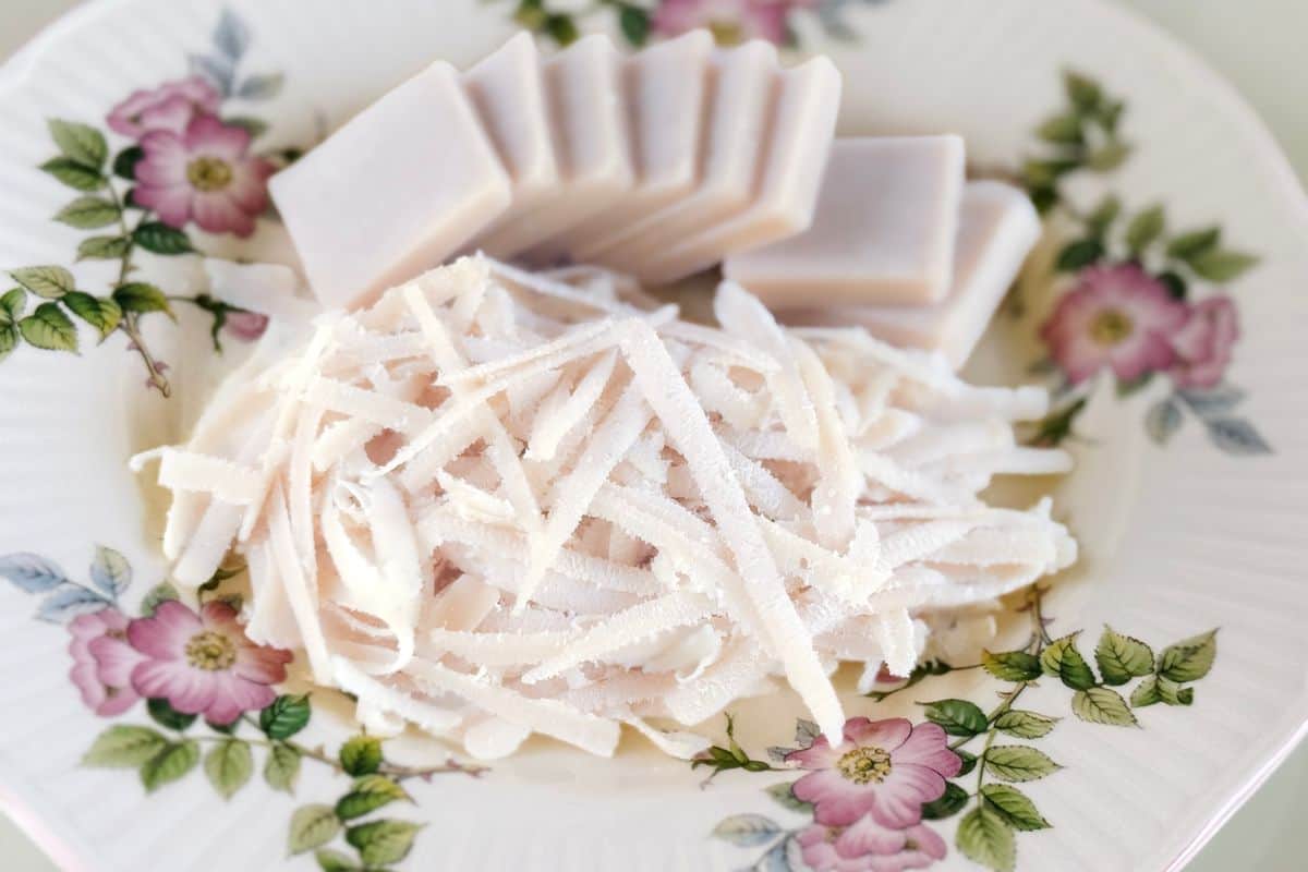 A block of homemade vegan mozzarella cheese sliced into pieces alongside a pile of freshly shredded vegan mozzarella, displayed on a vintage white floral plate with pink rose and green leaf details on a light grey surface.