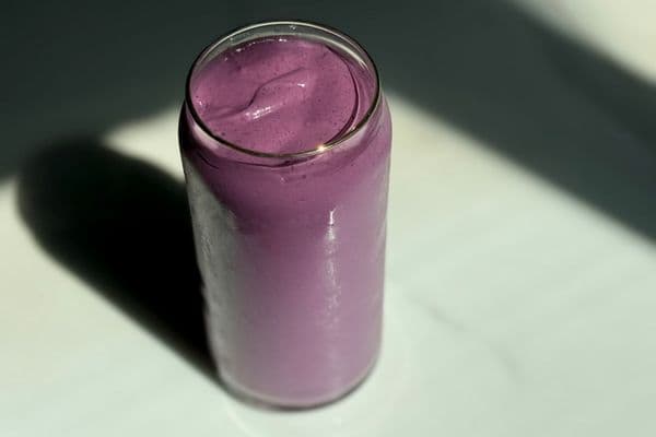 Overhead view of a creamy high protein plant-based peanut butter and jelly smoothie in a glass can cup on a dark marble surface with dramatic shadow lighting.