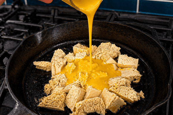 Golden vegan egg yolk sauce being poured over chunks of firm tofu in a cast iron skillet on a gas stovetop, in the process of making a plant-based tofu scramble.