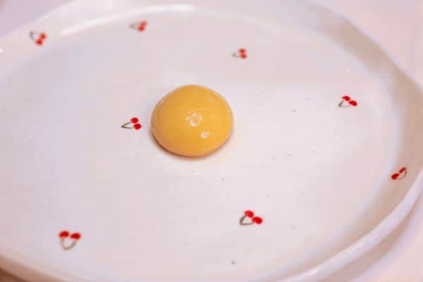 A single golden plant-based egg yolk sphere sitting on a white cherry patterned plate, created using spherification with Calcium Lactate and a Sodium Alginate bath.