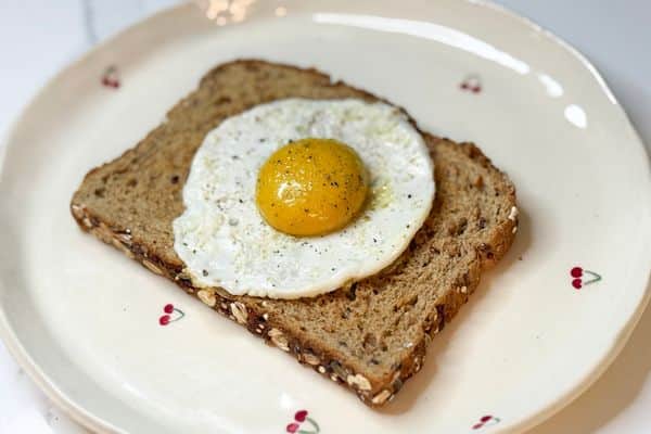 Plant-based fried egg on multigrain toast served on a white cherry plate, featuring a golden vegan egg yolk sauce on a vegan egg white, seasoned with black pepper.