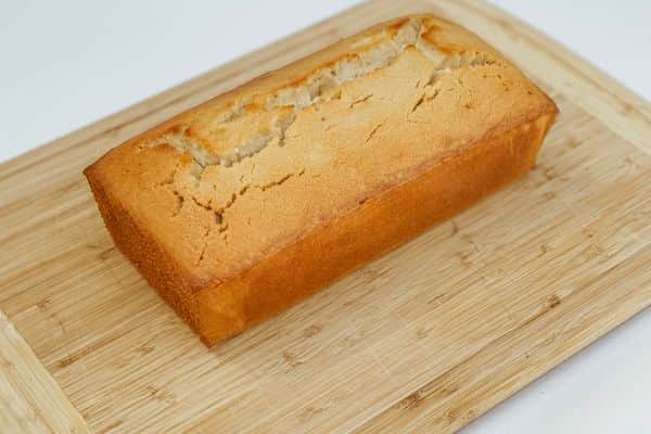 A golden-brown vegan pound cake with a perfectly cracked top crust, resting on a natural bamboo cutting board against a clean white background.