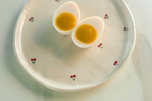 Two halves of a homemade vegan hard-boiled egg with a white tofu-based egg white and a golden plant-based yolk, displayed on a white ceramic plate with a small red cherry print pattern on a marble surface.