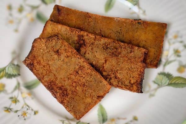 Three strips of crispy golden-brown tofu bacon with a deeply caramelized, smoky exterior, displayed on a vintage white ceramic plate with a delicate yellow floral and green leaf botanical pattern on a light background.