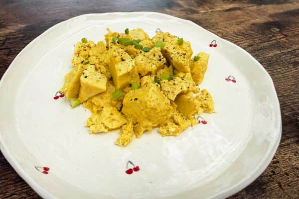 A golden plant-based tofu scramble on a white cherry patterned plate, seasoned with black pepper, plant-based egg sauce and topped with sliced green onions, served on a rustic wooden surface.