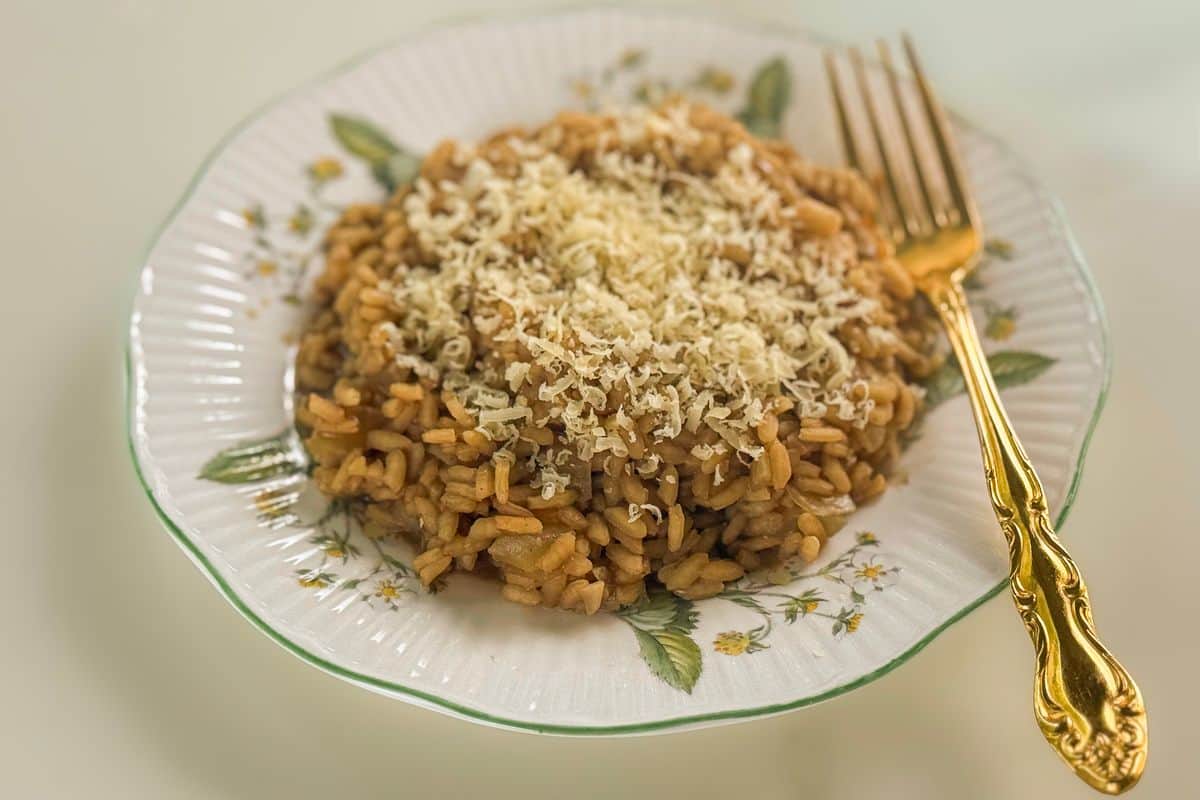 A generous serving of golden vegan truffle risotto topped with a snow of freshly grated vegan parmesan, plated on a vintage white ceramic plate with a green rim and floral botanical details, alongside an ornate gold fork on a neutral background.