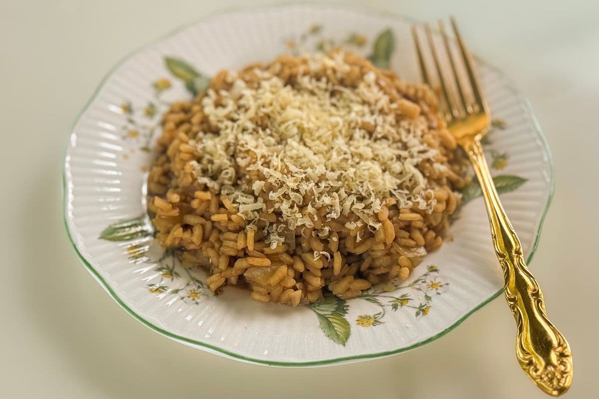 A generous serving of golden vegan truffle risotto topped with a snow of freshly grated vegan parmesan, plated on a vintage white ceramic plate with a green rim and floral botanical details, alongside an ornate gold fork on a neutral background.