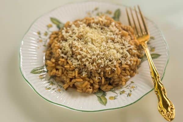 A generous serving of golden vegan truffle risotto topped with a snow of freshly grated vegan parmesan, plated on a vintage white ceramic plate with a green rim and floral botanical details, alongside an ornate gold fork on a neutral background.