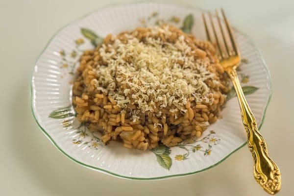 A generous serving of golden vegan truffle risotto topped with a snow of freshly grated vegan parmesan, plated on a vintage white ceramic plate with a green rim and floral botanical details, alongside an ornate gold fork on a neutral background.