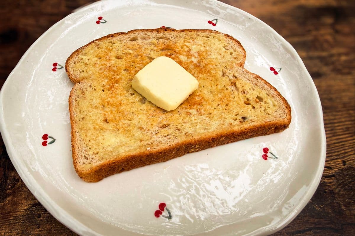 A slice of homemade plant-based butter melting on a slice of golden toasted multigrain bread on a white cherry patterned plate on a rustic wooden surface.