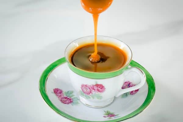A rich stream of golden-amber vegan caramel sauce being poured from a spoon into a vintage white and green floral teacup with pink rose details and gold trim, resting on a matching saucer against a white marble surface.