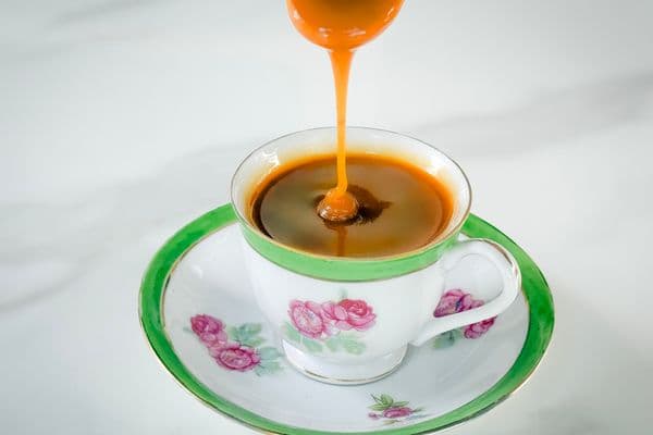 A rich stream of golden-amber vegan caramel sauce being poured from a spoon into a vintage white and green floral teacup with pink rose details and gold trim, resting on a matching saucer against a white marble surface.